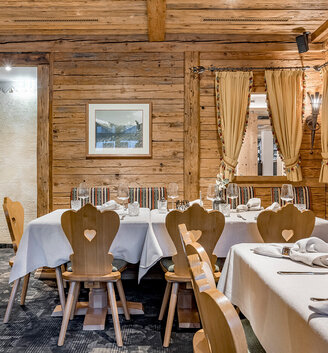 A cozy wooden restaurant interior featuring heart-shaped chairs, tables set with white tablecloths, curtains, framed artwork and warm lighting, creating an inviting, rustic atmosphere.