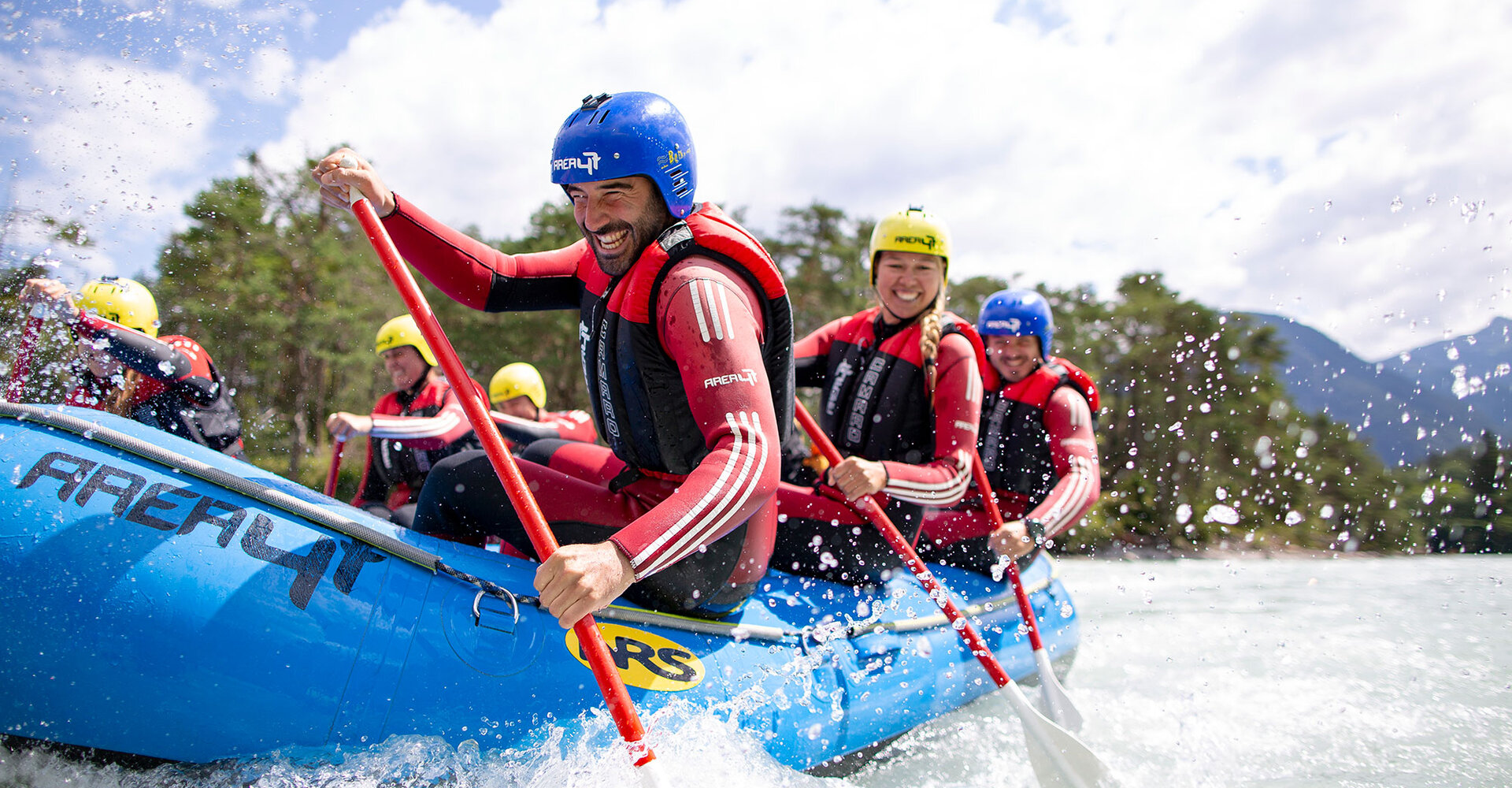 Eine Gruppe von Menschen, die Helme und Schwimmwesten tragen, paddelt in einem blauen Schlauchboot durch das spritzende Wasser, lächelt und genießt das Wildwasser-Rafting unter einem teilweise bewölkten Himmel.