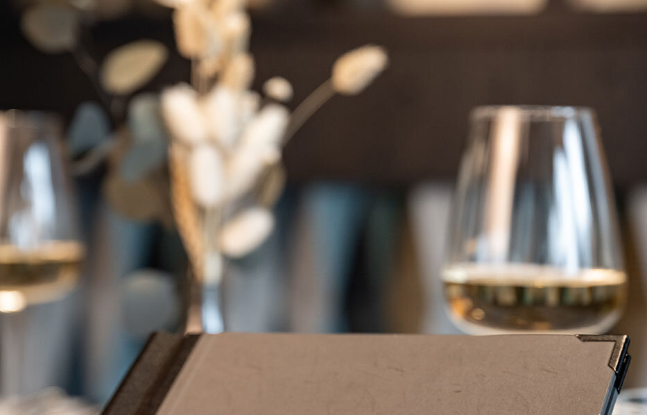 A person is holding a menu and sitting at a restaurant table with two glasses of white wine and a vase of dried flowers while the background is blurred.