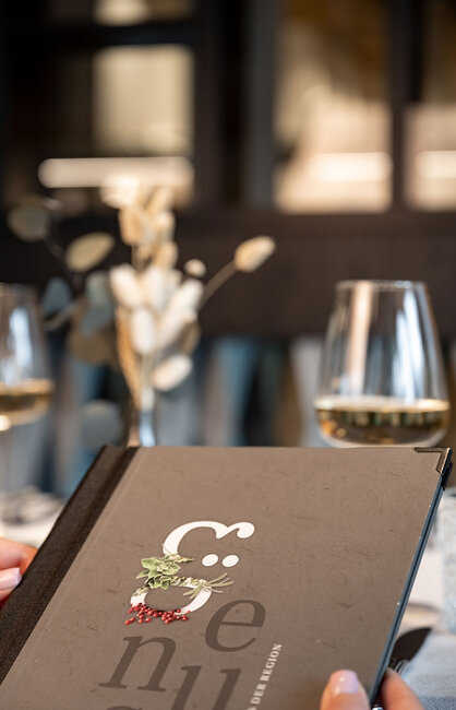 Eine Person hält eine Speisekarte in der Hand und sitzt an einem Restauranttisch, auf dem zwei Gläser Weißwein und eine Vase mit Trockenblumen stehen; der Hintergrund ist unscharf.