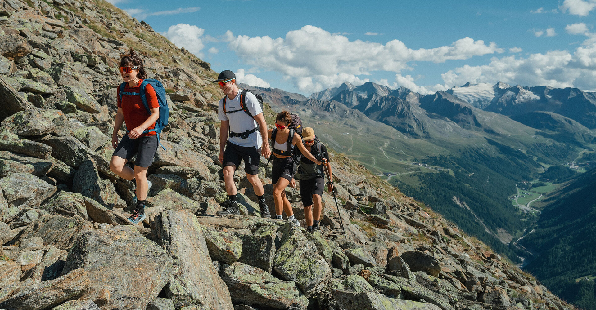Four hikers with backpacks climb a rocky mountainside under a bright blue sky, surrounded by distant peaks and a green valley below them.