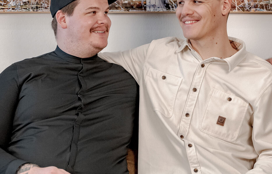 Two men are sitting close together on a sofa, smiling at each other. One is wearing a black shirt and a cap, the other a white button-down shirt. A large painting of an animal hangs on the wall behind them.