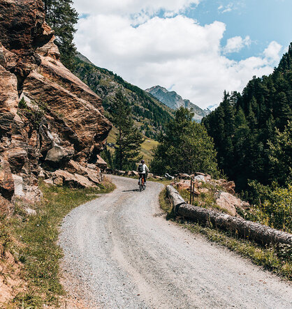A cyclist rides along a winding gravel path through a picturesque mountain landscape, surrounded by rocky cliffs, green trees and distant peaks under a partly cloudy sky.