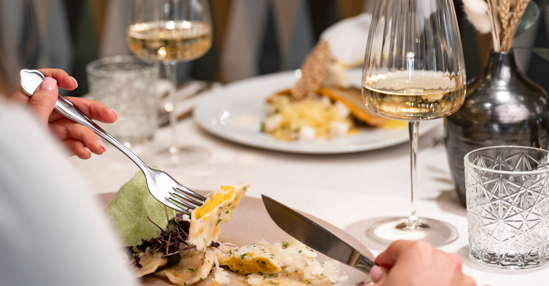 An attractively presented gourmet dish of fish on risotto, garnished with a green crisp and a wrapped lemon, is placed on a white tablecloth next to two wine glasses in an elegant restaurant.