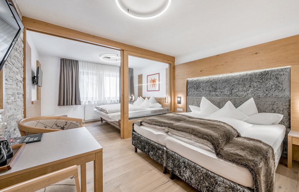 Modernes Hotelzimmer mit zwei großen Betten, hellem Holzboden, einer Akzentwand aus Stein, einem Schreibtisch, einem Fernseher und einem großen Fenster mit weißen Vorhängen. Das Zimmer ist in neutralen Farben gehalten und mit eleganten, modernen Möbeln eingerichtet.
