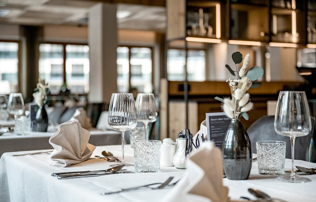 An elegantly set restaurant table with wine glasses, folded napkins, cutlery and a small vase of dried flowers. Shelves and large windows are in the background, creating a modern, inviting atmosphere.