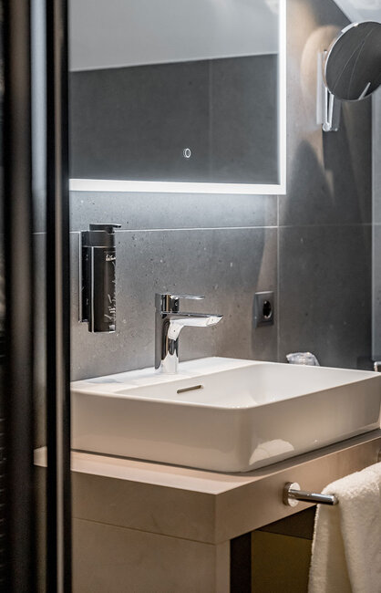 Modernes Badezimmer mit einem rechteckigen weißen Waschbecken, einer verchromten Armatur, einem großen Spiegel mit LED-Beleuchtung, einer dunkelgrauen Fliesenwand, einem Handtuchhalter und an der Wand montierten Accessoires.
