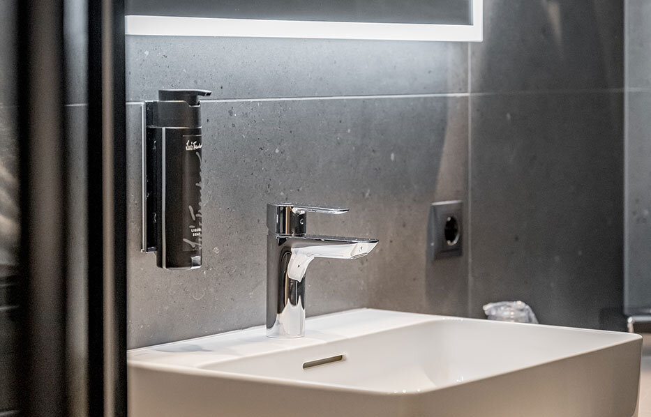 Modern bathroom with a rectangular white washbasin, a chrome faucet, a large LED-lit mirror, a dark gray tiled wall, a towel holder and wall-mounted accessories.