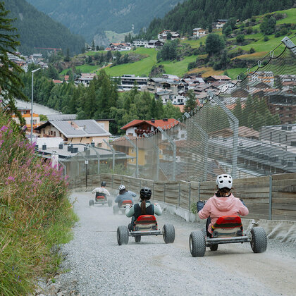 People wearing helmets ride mountain carts along a gravel path under a cloudy sky, surrounded by mountains, trees and alpine village houses.