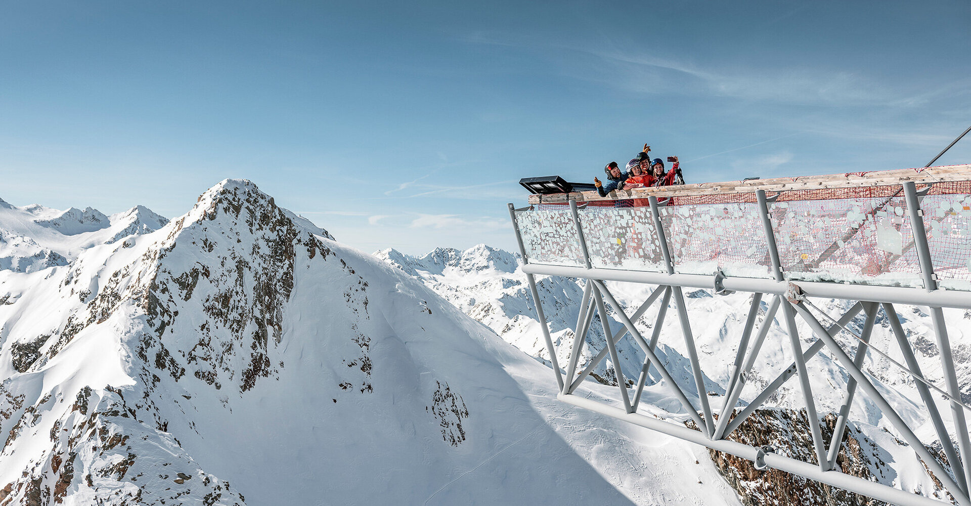A group of people standing on a viewing platform made of steel above snow-covered mountains under a bright blue sky, admiring the picturesque Alpine landscape.