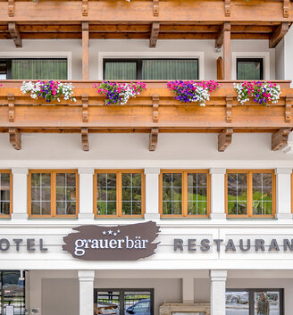 The façade of Hotel "Grauer Bär" features balconies adorned with colorful flower boxes and large windows. The building is designed in a rustic alpine style, featuring wood paneling and white walls.