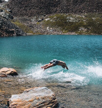 An einem sonnigen Tag springt jemand kopfüber in einen klaren, blauen See, der von Felsen und Bergen umgeben ist.
