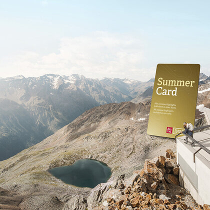 Two people stand on a platform next to a huge "Summer Card" sign, looking out at a small lake and rugged mountain peaks under a clear sky.
