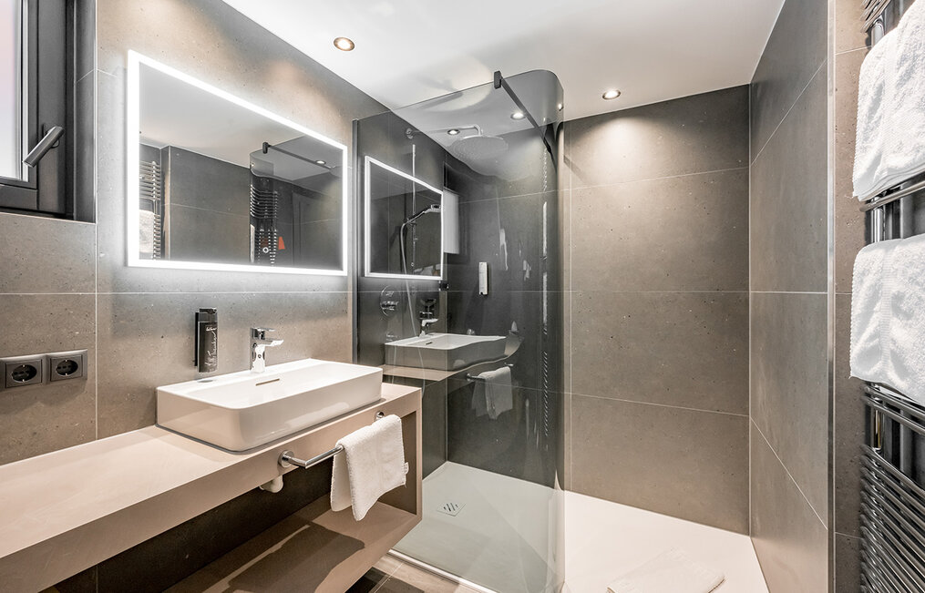Modern bathroom with a large mirror, rectangular washbasin, cantilevered vanity top, walk-in glass shower, gray-tiled walls, towel holder and recessed ceiling lights.