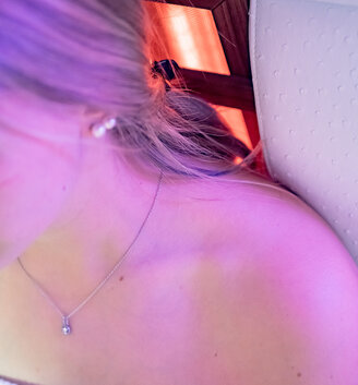 A woman with blonde hair and a necklace is lying under violet light on an infrared bench, wearing a white, textured towel. Only her shoulder, neck and part of her face are in the picture.
