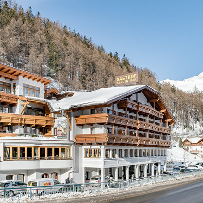 A large hotel with wooden balconies stands next to a snow-covered road, surrounded by trees and mountains under a brilliant blue sky. Cars are parked along the road.