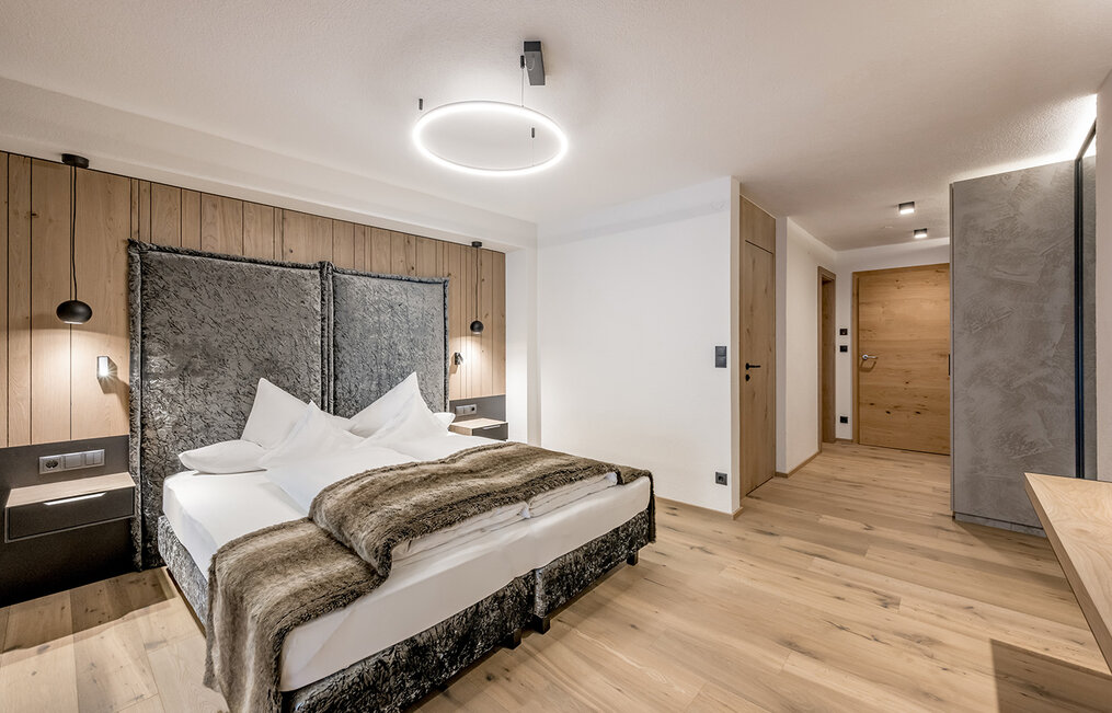 Modern bedroom featuring a double bed, a fur throw, wooden floors, a round ceiling light, wooden wall paneling, nightstands and a wooden door in the background. The room has a minimalist yet cozy design.