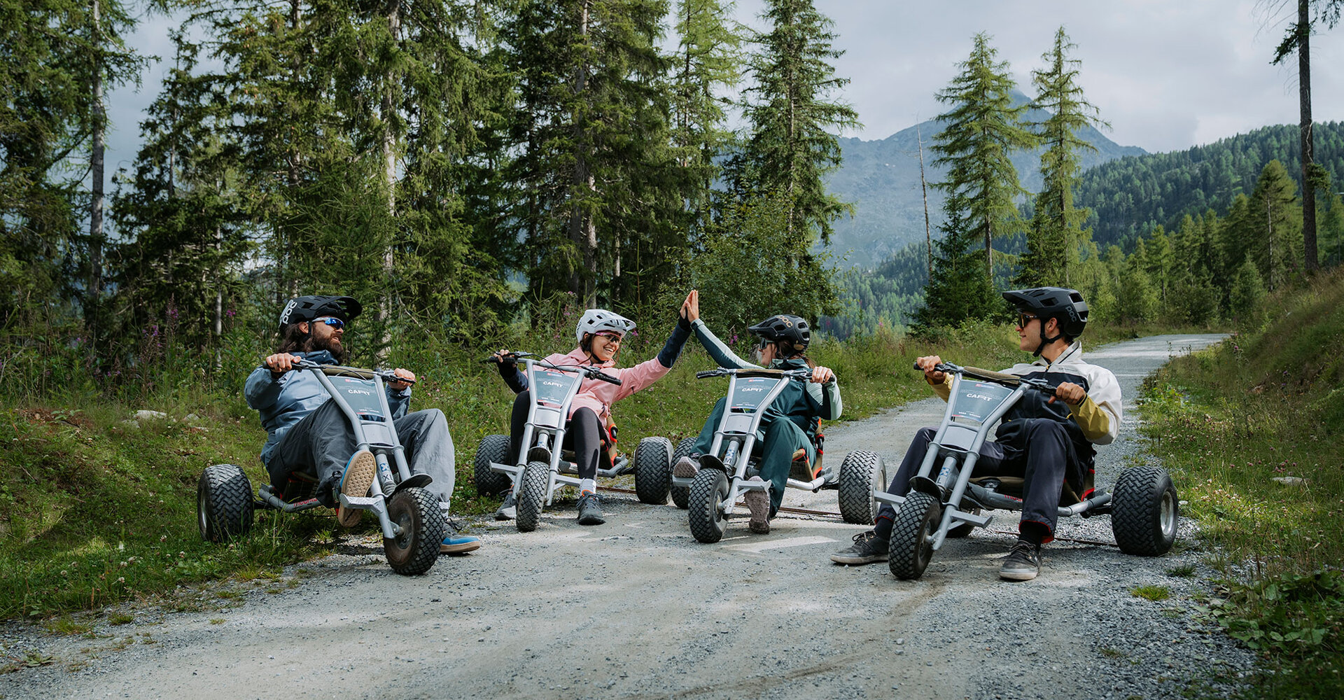 Four people wearing helmets are riding tricycles on a gravel path through a wooded area. Two of them give each other a high-five. Pine trees and mountains are in the background.