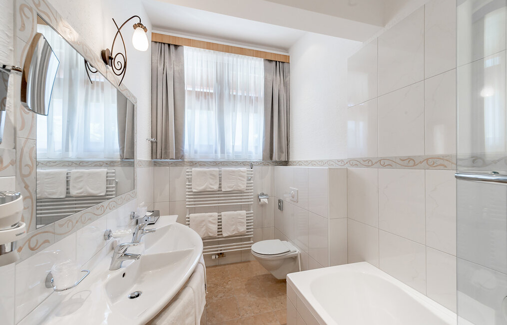Bright bathroom with white tiles, a double sink, a bathtub, a wall-mounted toilet, a large mirror, towel racks with folded towels and two curtained windows that let in daylight.