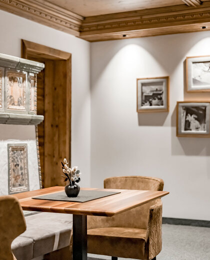In a cozy corner stands a small table with two light-brown chairs. Framed black-and-white photos hang on the white wall and a vase of dried flowers stands on the table. The warm lighting creates an inviting atmosphere.