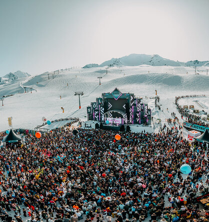 A large crowd gathers at an open-air music festival on a snow-covered mountain, featuring a stage, colorful lights and performing DJs. Snow-capped peaks and ski lifts are in the background.