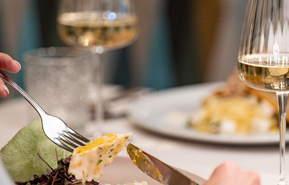 A person is cutting food on a plate at a restaurant table. In the background, there are two glasses of white wine and another plate of food.
