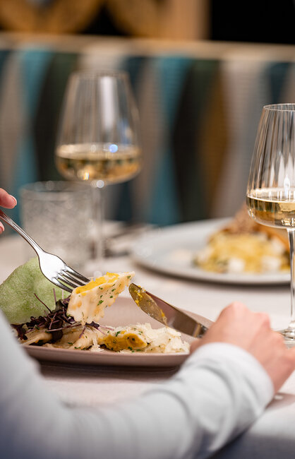 Eine Person schneidet an einem Restauranttisch Essen auf einem Teller, im Hintergrund stehen zwei Gläser Weißwein und ein weiterer Teller mit Essen.