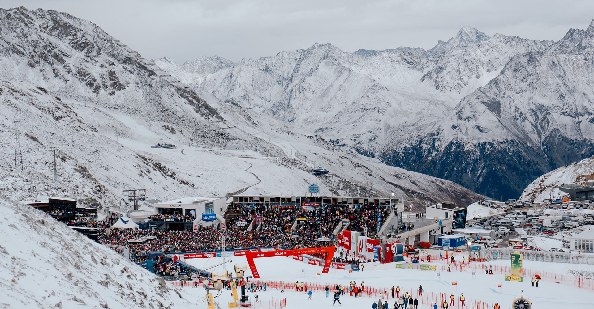 A snow-covered mountain landscape where a large crowd has gathered near ski gates and grandstands for a ski race. Snow-capped peaks and an overcast sky form the backdrop.