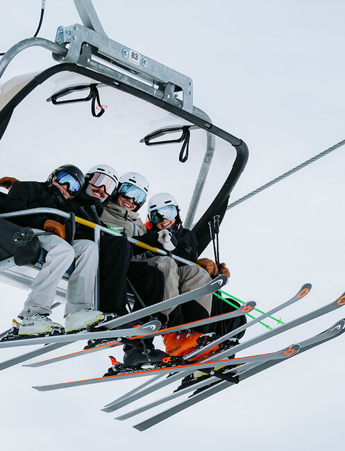 Four people wearing helmets and ski gear are riding uphill on a ski lift, holding ski poles and their skis and snowboards against a backdrop of a snowy, cloudy sky.