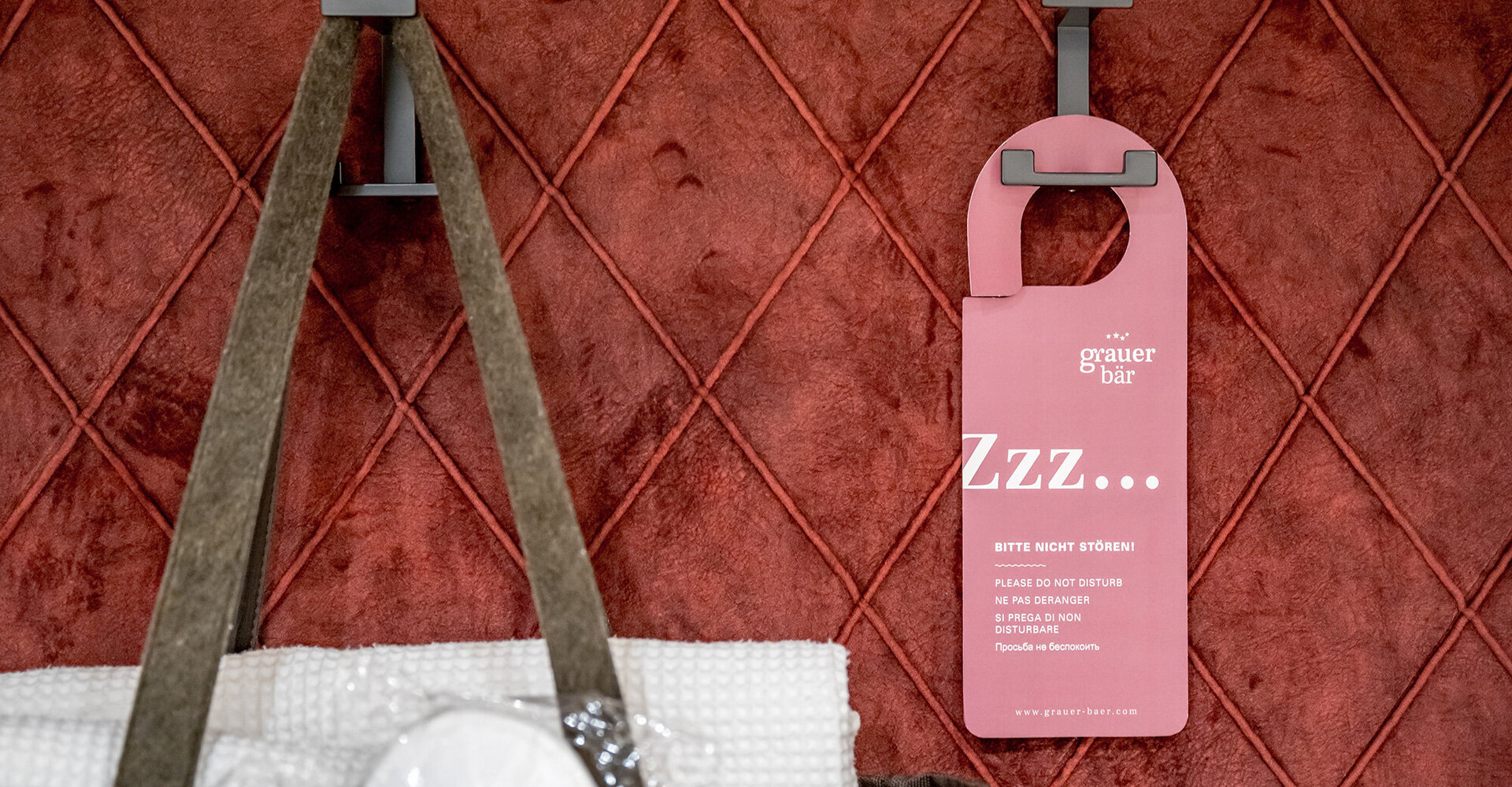A pink door hanger and a bag containing towels hang on hooks in front of a red quilted wall.