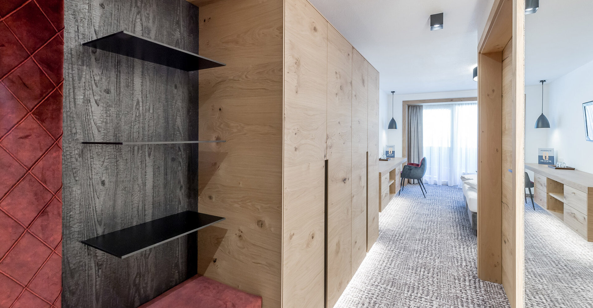 A modern hotel room with light wood paneling, floating black shelves, an upholstered red bench, a textured carpet, a desk, a chair, a large window and minimalist furnishings. The room is bright and spacious.