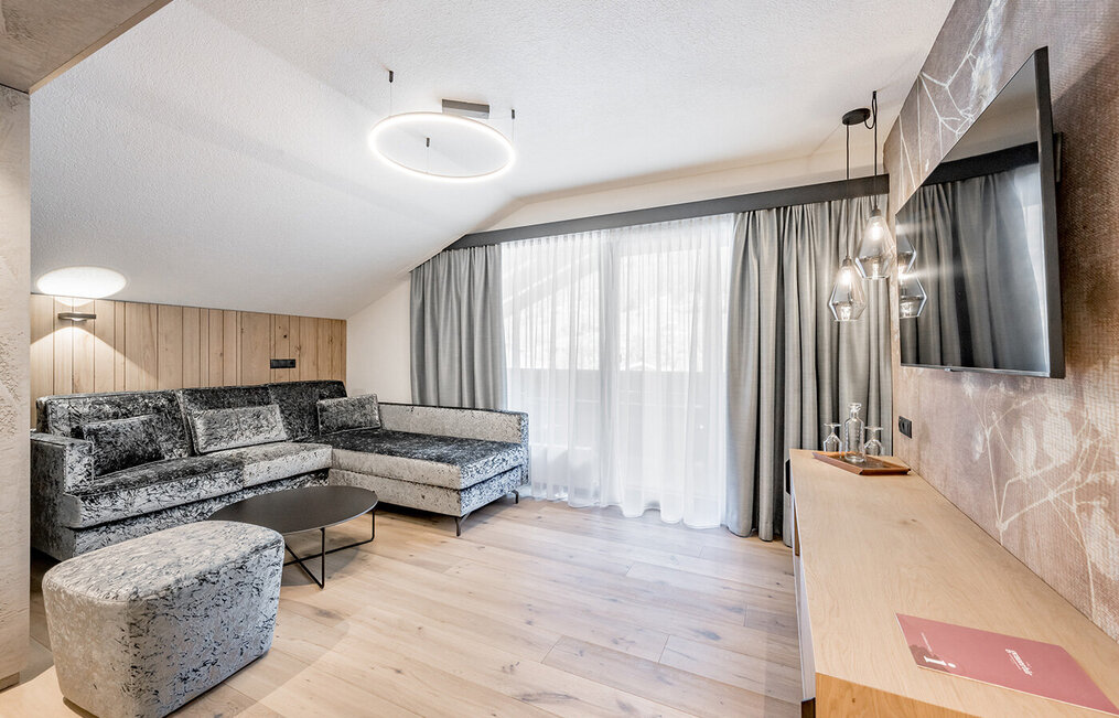 Modern living room with a silver sofa, a round black coffee table, a wall-mounted TV, wooden flooring, long curtains and modern lighting fixtures. Light streams in through the large windows.