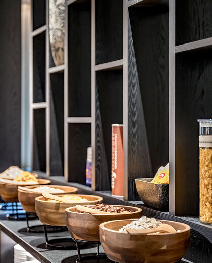 Wooden bowls filled with various breakfast cereals sit on a buffet counter with shelves and decorative items in the background.