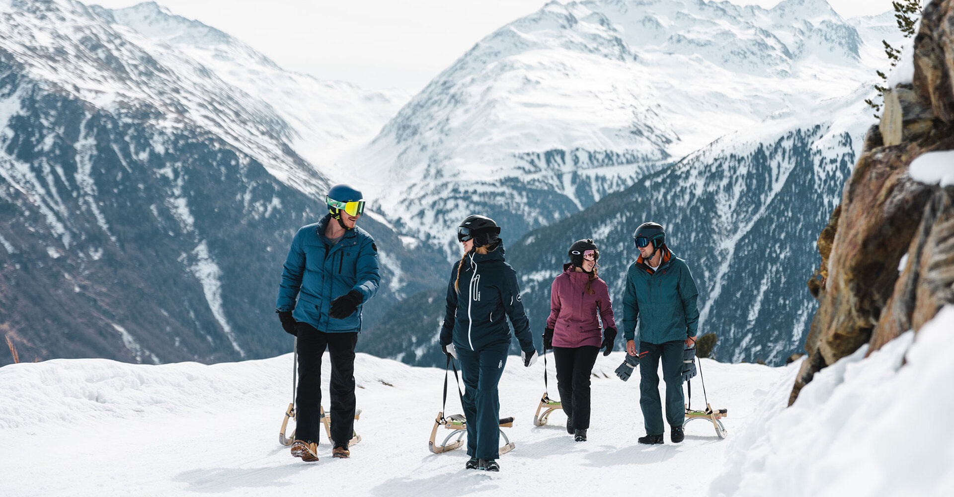 Four people in winter clothing are walking with toboggans on a snowy mountain path. Snow-covered peaks are in the background.