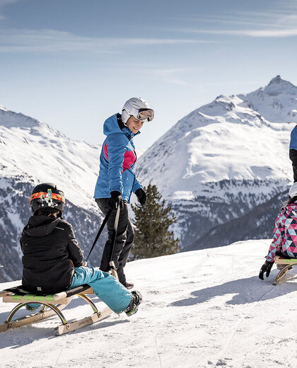 Four people, two adults and two children, wearing helmets and winter clothing are on a snow-covered mountain with sleds. Snow-capped peaks and a clear sky in the background.