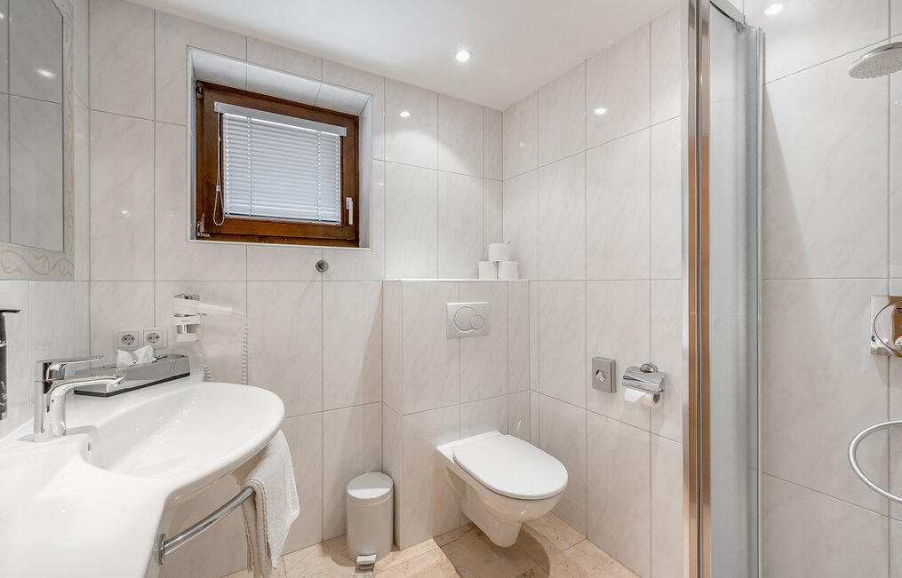 Modern, white-tiled bathroom featuring a wall-mounted toilet, a washbasin, a shower with a glass door, a small window with blinds and a waste bin. Bright lighting and furnishings create a clean, uncluttered look.