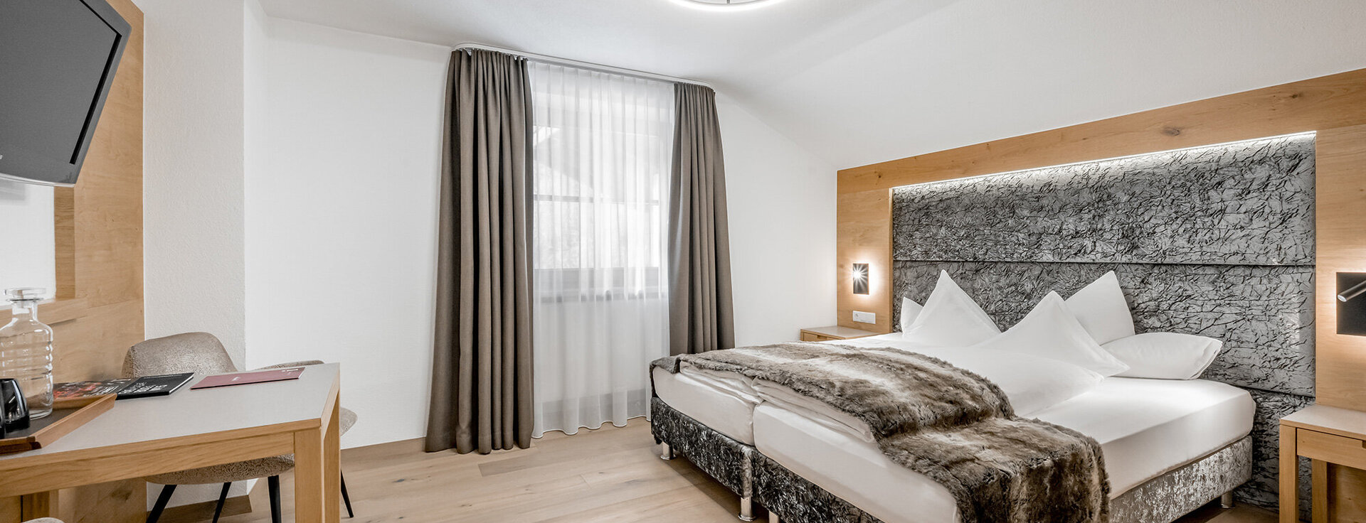 Modern hotel room with a large bed, wooden flooring, a desk with a chair, a wall-mounted TV, a window with sheer curtains and neutral décor.