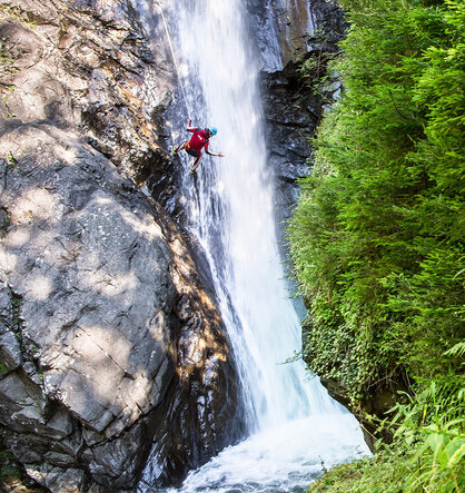 A person wearing a helmet and red clothing is rappelling down a waterfall next to a rocky cliff, surrounded by green foliage.