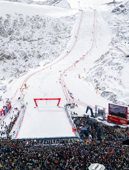 At the foot of a snow-covered ski slope, a large crowd gathers for a ski race, the course of which is marked by red gates and winds its way down the steep mountain.