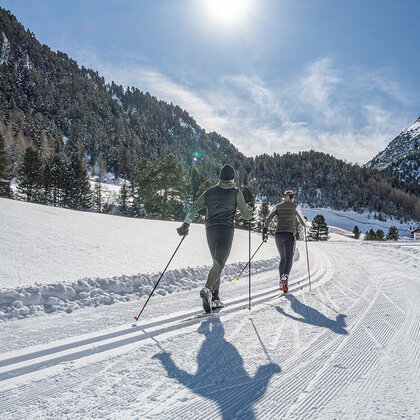Two people are skiing along a groomed trail through a sunny mountain landscape, surrounded by trees and a brilliant blue sky.