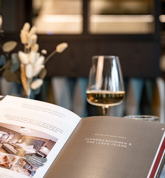 A close-up of an open menu on a table, featuring a wine glass and a vase of dried flowers, in a softly lit, modern restaurant.