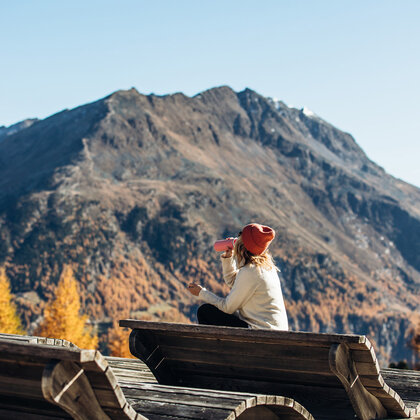 A person wearing a red hat sits on a wooden deck chair and drinks from a bottle. In the background, therer are autumn trees and a large mountain under a brilliant blue sky.