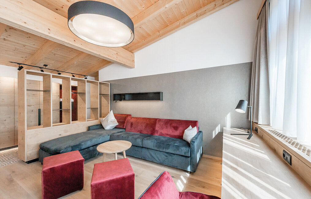 Modern living room with a wooden ceiling, a large gray corner sofa with red cushions, two red stools, a round wooden coffee table and sunlight streaming in through tall windows with light-colored curtains.