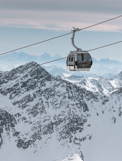 A mountain gondola cabin travels over snow-capped mountain peaks with rugged slopes, set against a cloudy sky with distant mountain ranges in the background.