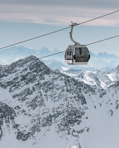 A mountain gondola cabin travels over snow-capped mountain peaks with rugged slopes, set against a cloudy sky with distant mountain ranges in the background.