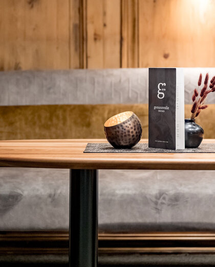 In front of a grey sofa with a gold cushion and a wood-paneled wall in the background stands a wooden table holding a dark vase, a round wooden bowl and a drinks menu.