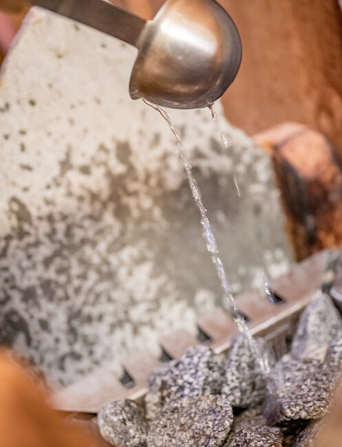 Aus einer Metallkelle wird Wasser auf heiße Saunasteine gegossen, wodurch Dampf entsteht. Im Hintergrund ist das Innere einer Sauna mit Holzelementen zu sehen.
