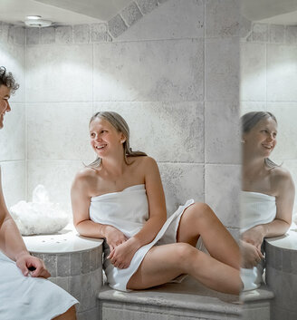 A man and a woman, clad only in towels, are sitting in a white-tiled steam room, chatting and smiling at each other. Behind them stands a decorative bowl containing crystals.