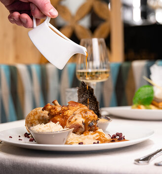 A hand pours sauce over a dish featuring a pork knuckle served with rice and vegetables. Next to it, a glass of red wine stands on a white tablecloth in a restaurant.