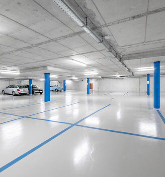 Underground garage with a polished floor, blue parking space markings, blue columns and a few parked cars. The area is brightly lit by fluorescent lights.